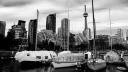 Black and white photo of sailboats docked at a marina, some under white winter covers, with high-rise buildings and the CN Tower in the background.
